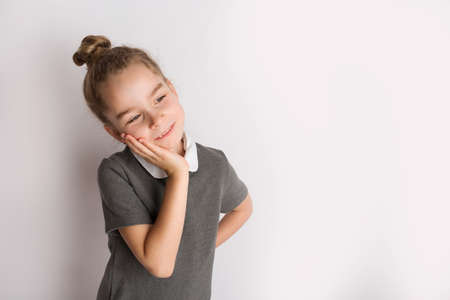 Attractive little girl in a strict school dress, stands on a white background with empty copy space, gestures with her hands, admires a happy expression on her faceの写真素材
