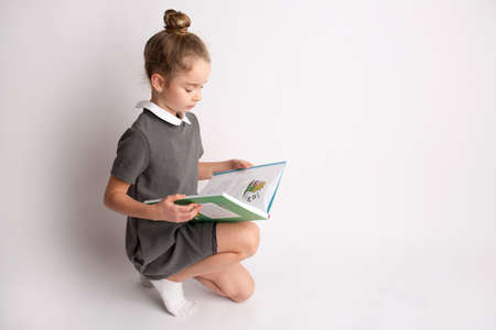 Attractive little girl in a strict school dress, stands on a white background with empty copy space, gestures with her hands, admires a happy expression on her faceの写真素材