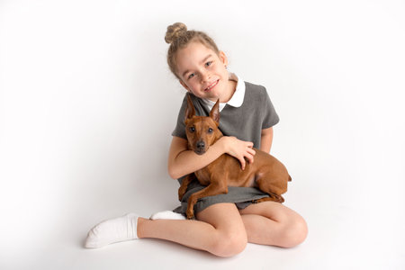 Attractive little girl in a strict school dress with a dwarf brown pinscher on a white background admires a happy expression on her faceの写真素材