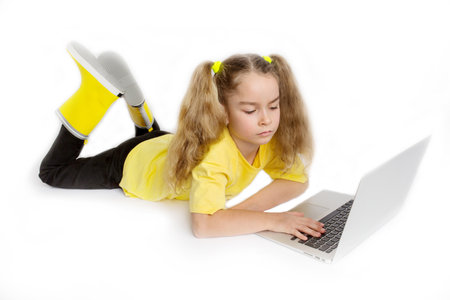 Concentrated little cute girl child yellow t-shirt lying in front of computer, checking tasks in online classroom, distance learning concept.の写真素材