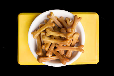 Close-up of brown dog food bones in a white dog bowl on a black studio background. Yellow bowl holder.の写真素材