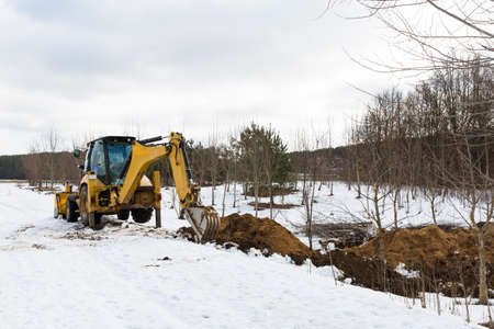 A yellow tractor excavator digs the ground for the repair and laying of underground utilities and cables for electricity and communications. During the winter season.の写真素材