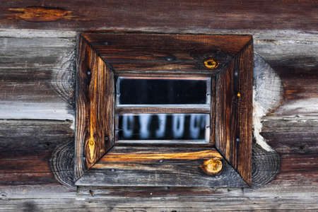 Rustic wooden house made of round logs. Ancient window design. Wooden window frame. Snow covered...