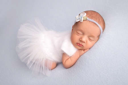 Sleeping newborn girl in the first days of life in a white ballet dress with a white bandage and a blue flower on a blue background. Studio macro photography, portrait of a newborn ballerina.の写真素材