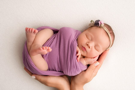 Sleeping newborn girl in the first days of life on a white background. A newborn baby in a purple lilac winding and a headband. Hand, palms of father and mother, parents hold the child.の写真素材