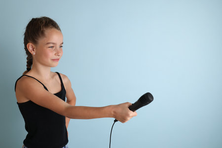 Portrait of a cute teenage girl singing, using microphone. Teenage girl in black t-shirt on an isolated blue background. Karaoke for kids, home entertainment for kids. Singer singing with microphone.の写真素材