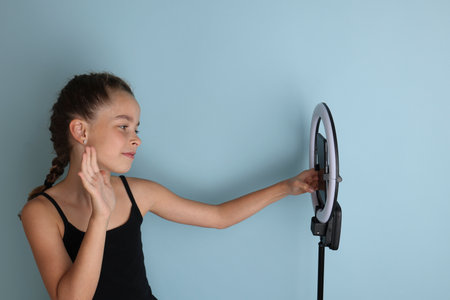Little emotional teenage girl in a black t-shirt on an isolated blue background. Studio portrait of a young smiling teenage girl, vlogger, with a ring flash lamp and a smartphone. Girl takes a selfie.の写真素材