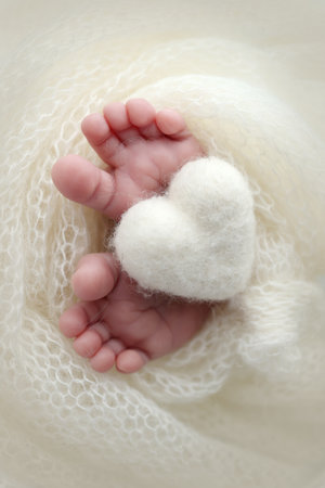 Knitted white heart in the legs of a baby. Macro photography. The tiny foot of a newborn baby. Soft feet of a new born in a white wool blanket. Close up of toes, heels and feet of a newborn.の写真素材