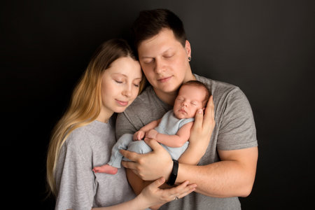 A cute newborn baby boy sleeps in a blue overalls in the first days of life. In the arms of father and mother. Warm parental hugs. On a black background. Professional portrait photography.の写真素材