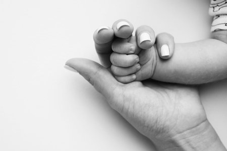Parents hands hold the fingers of a newborn baby. The hand of a mother and father close-up holds the fist of a newborn baby. Family health and medical care. Tiny fingers in a black and white photo.の写真素材