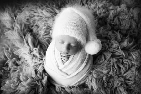 A sleeping newborn boy in the first days of life in a white soft cocoon with a knitted woolen white hat on a white background in the shape of a heart. Black white photography, portrait of a newborn.の写真素材