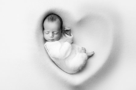 A sleeping newborn boy in the first days of life in a white soft cocoon with a knitted woolen white hat on a white background in the shape of a heart. Black white photography, portrait of a newborn.の写真素材