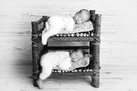 Tiny newborn twin boys in white bodysuits against a light wood background. Newborn twins sleep on a...