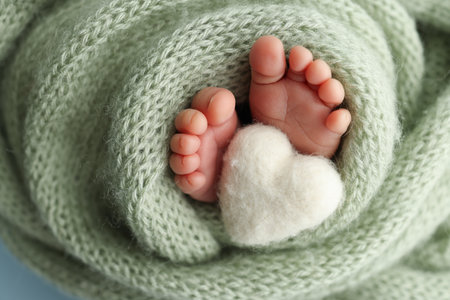 The tiny foot of a newborn baby. Soft feet of a new born in a olive green wool blanket. Close up of toes, heels and feet of a newborn. Knitted white heart in the legs of a baby. Macro photography.の写真素材