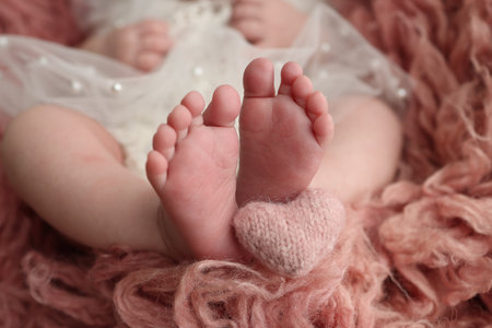 The tiny foot of a newborn baby. Soft feet of a new born in a pink red flakatti. Close up of toes, heels and feet of a newborn. Knitted pink red heart in the legs of a baby. Openwork white dressの写真素材