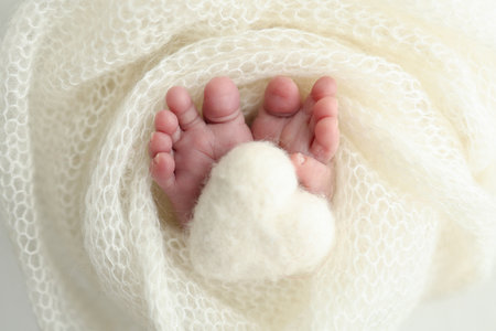 Soft feet of a new born in a white wool blanket. Knitted white heart in the legs of a baby.