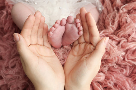 Mother holding the feet of a newborn baby on a background of pink flakatti. Feet of a newborn in the hands of parents. Macro photo of the foot, heels and toes. Openwork white dress Parents palms.の写真素材