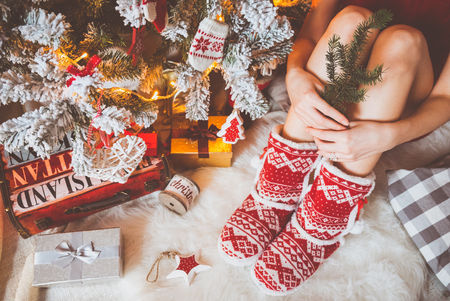 Young pretty woman in warm cozy clothes is sitting on the light wooden floor in her bright house near the christmas tree and holds a branch of fir in her hands.の写真素材