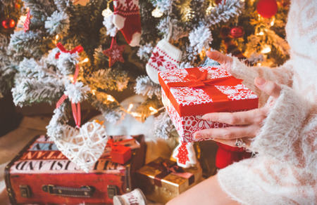 Young pretty woman in warm cozy clothes is sitting on the floor in her bright house near the christmas tree and holds gift box in her hands.の写真素材