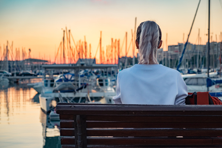Young pretty girl is sitting on a bench and looking at the bay with yachts at sunsetの写真素材