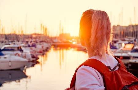 Young pretty girl is standing on a pier and looking at the bay with yachts at sunsetの写真素材