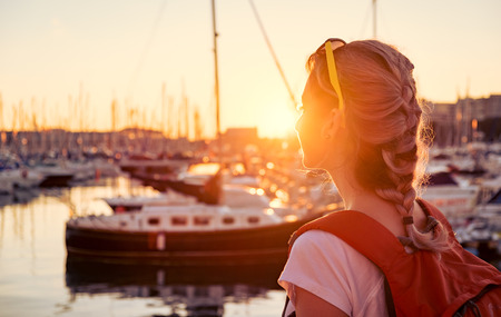 Young pretty girl is standing on a pier and looking at the bay with yachts at sunsetの写真素材