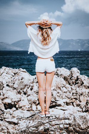 Beauty tanned and stylish young woman in white clothes and straw hat enjoys standing on stone shore at the seaside in sunny summer day and looking away. Travel and vacation concept.の写真素材