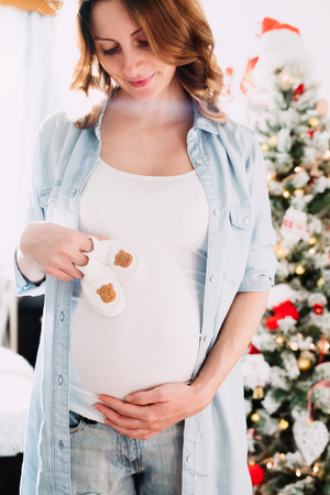 Happy beautiful pregnant woman holds Cute little infants shoes in hands at christmas tree in bright white room and touching her belly.の写真素材