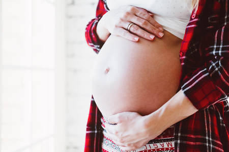 Happy beautiful pregnant woman standing at the window in bright white room and touching her belly.の写真素材