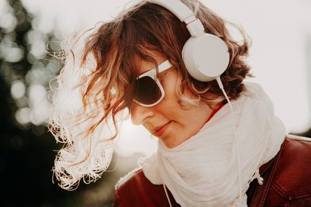 Beautiful young woman in a sunglasses and headphones listening to music against bright sunset....