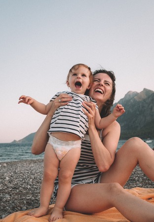 Young mother with her little baby girl have fun sitting on a beach in summer day at sunset over mountains. Happy family concept. Happy childhood.の写真素材