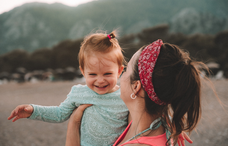 Young mother with her little baby girl have fun sitting on a beach in summer day at sunset over mountains. Happy family concept. Happy childhood.の写真素材