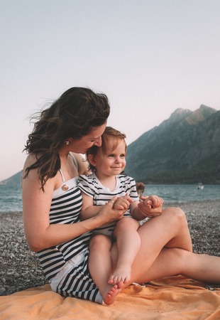 Young mother with her little baby girl have fun sitting on a beach in summer day at sunset over mountains. Happy family concept. Happy childhood.の写真素材