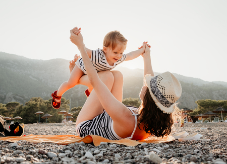 Young mother with her little baby girl have fun sitting on a beach in summer day at sunset over mountains. Happy family concept. Happy childhood.の写真素材