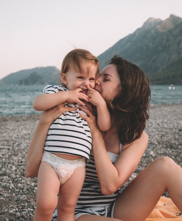 Young mother with her little baby girl have fun sitting on a beach in summer day at sunset over mountains. Happy family concept. Happy childhood.の写真素材