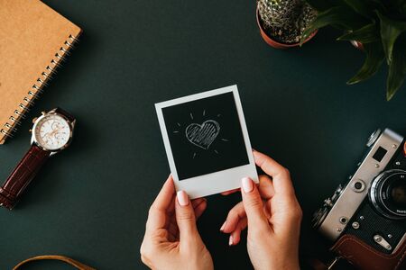 Top view flat lay female hand holds  picture with heart next to film camera in case wristwatch and notebook lie on dark tabletop. Concept of memories of a romantic trip. Advertising spaceの写真素材