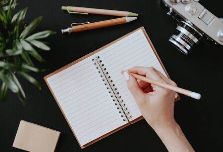 Top view of a flat lay female hand writes notes in an open notebook lying on a black table next to colored pens with paper and a film camera. Diary concept. Advertising spaceの写真素材