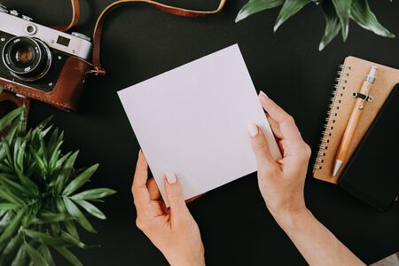 Top view of flat lay female hands holding white sheet of page next to film camera in case notebook smartphone and pen on black table. Workspace concept of a creative person. Advertising spaceの写真素材