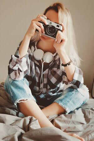 Young beauty female in casual shirt taking a picture with her vintage camera sitting on a bedの写真素材