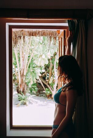 Happy young beautiful female stands at opened window in her bungalow and looks outside to tropical garden. Travel and vacation conceptの写真素材