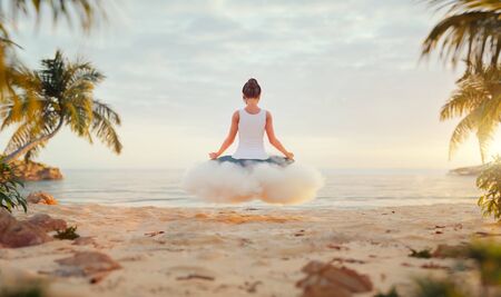 3d illustration of a Young Happy Carefree Woman Enjoying Beautiful Sunset sitting on the white cloud in lotus position on the Tropical Beach. Yoga and meditation concept.の写真素材