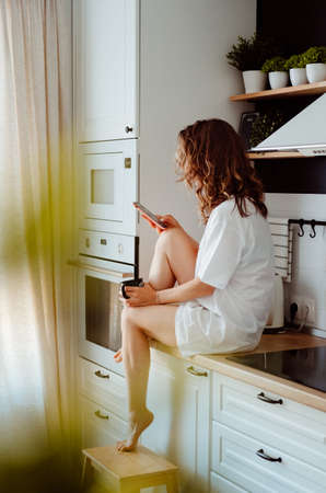 Young pretty woman drinks coffee sitting at her kitchen at home and using smartphone enjoys of morning. Smiling woman reading phone messageの写真素材