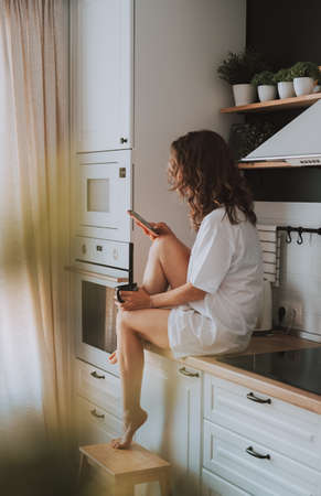 Young pretty woman drinks coffee sitting at her kitchen at home and using smartphone enjoys of morning. Smiling woman reading phone message. Vintage film lookの写真素材