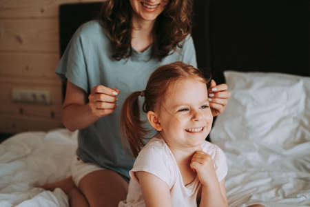 Mother is combing her daughter's hair sitting on the bed in the room. Happy kid and mom have fun.の写真素材