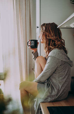 Young pretty woman drinks coffee sitting at her kitchen at home and looking out the window enjoys of morning. Vintage film lookの写真素材