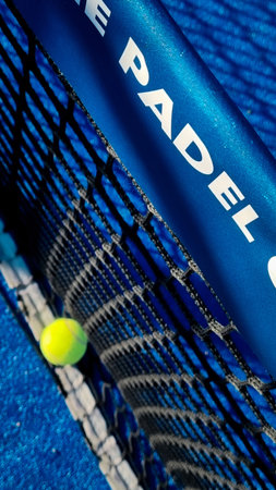 Close-up of a yellow tennis ball resting against a black net on a blue padel court, with part of the net post visible.の写真素材