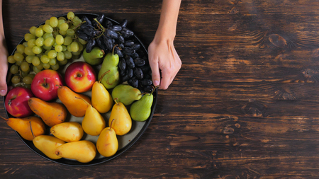 Unrecognizable woman holding tray different season fruit over the wooden table. Close up female hands with grapes, pears, applesの写真素材