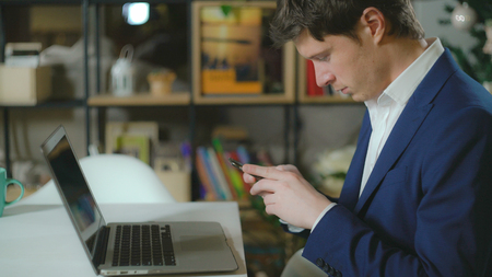 Handsome man sitting in room with bookshelves on the desk laptop. Elegant fashionable man wearing in casual blue suit texting message on smartphone.の写真素材