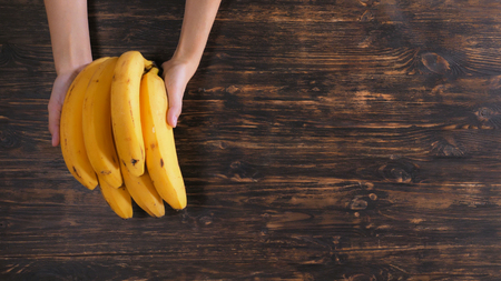 Unrecognizable woman holding fruits over the wooden table. Close up female hands with bananaの写真素材