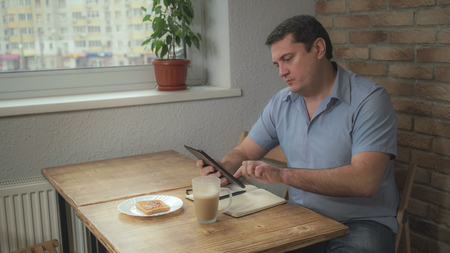 Adult male in a blue shirt in the morning sitting in the hotel drinking coffee and eating toast, is on the table and tablet daily, Soft panoramaの写真素材
